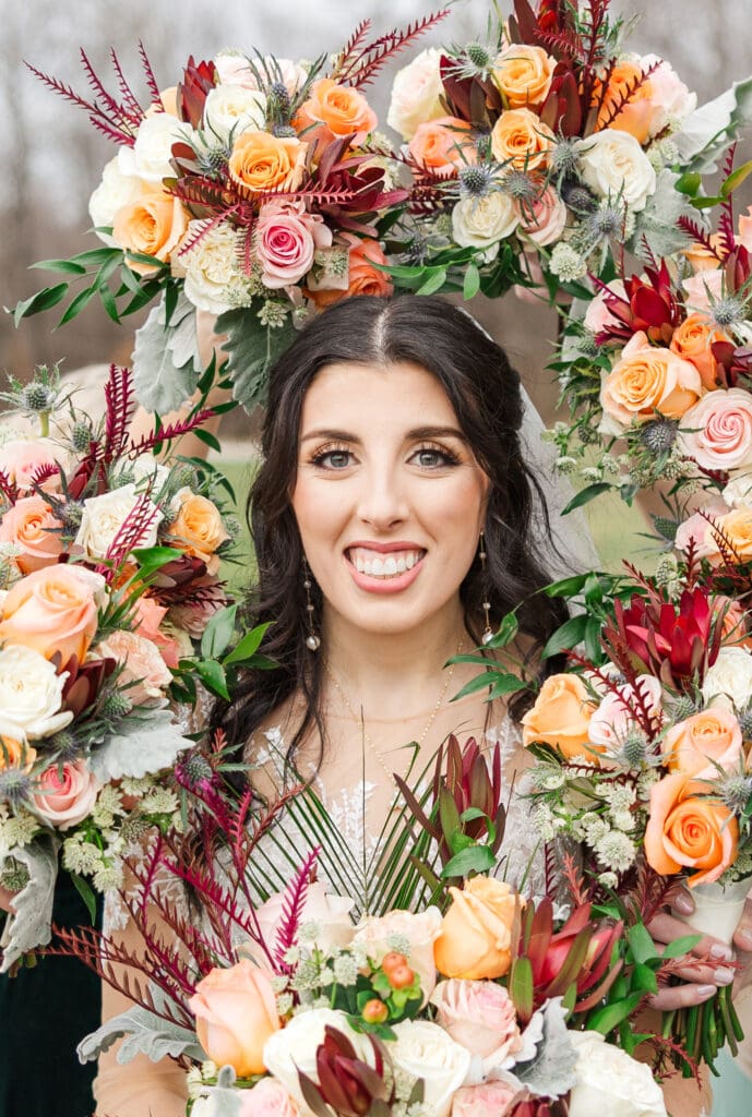 Bride at a winter wedding at Hidden Homestead in Smiths Grove, Kentucky, framed by lush green foliage and warm peach and ivory florals, smiling softly outdoors